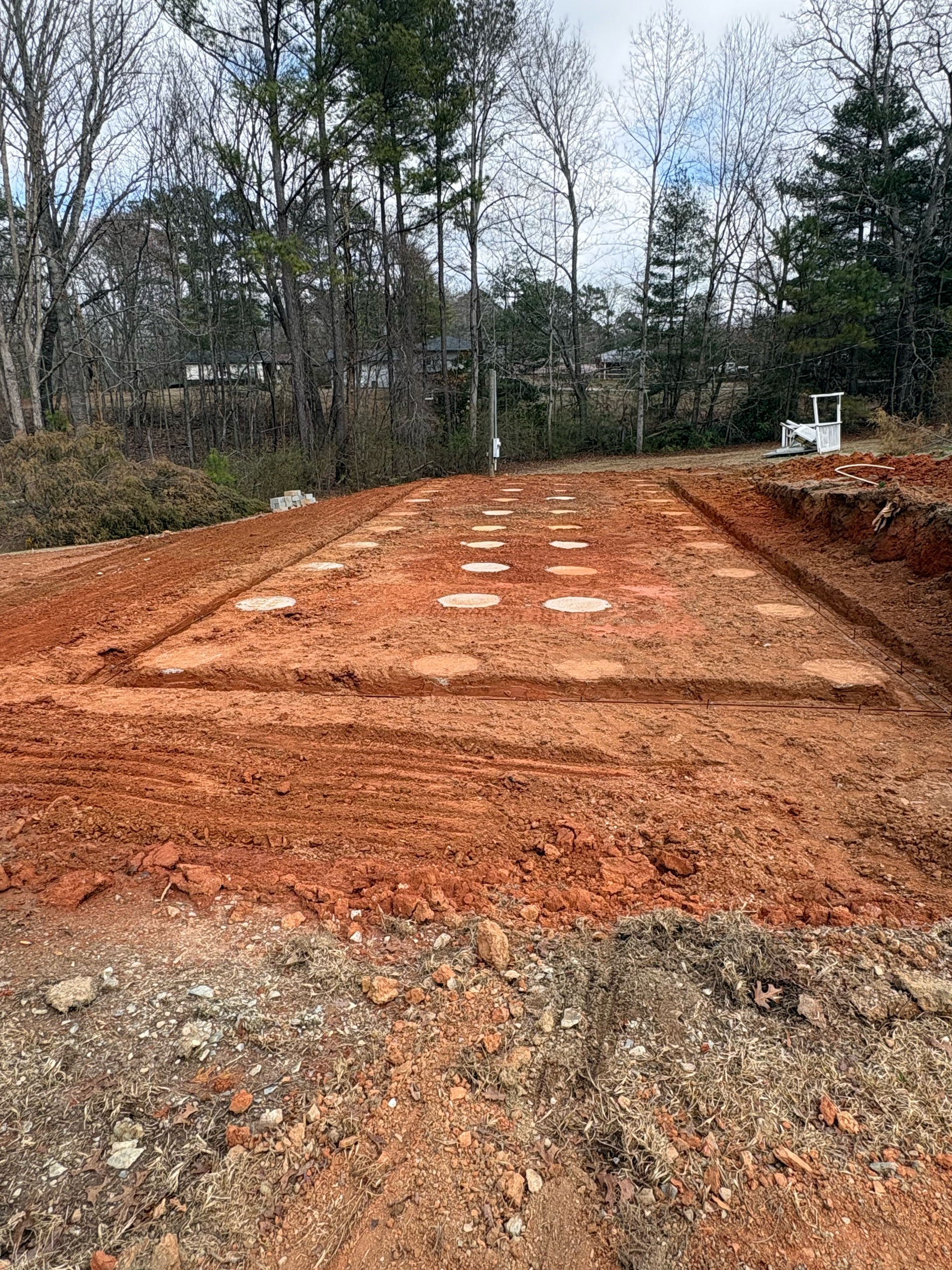 A construction site with a grid of circular concrete footings embedded in red clay, viewed from an elevated perspective.