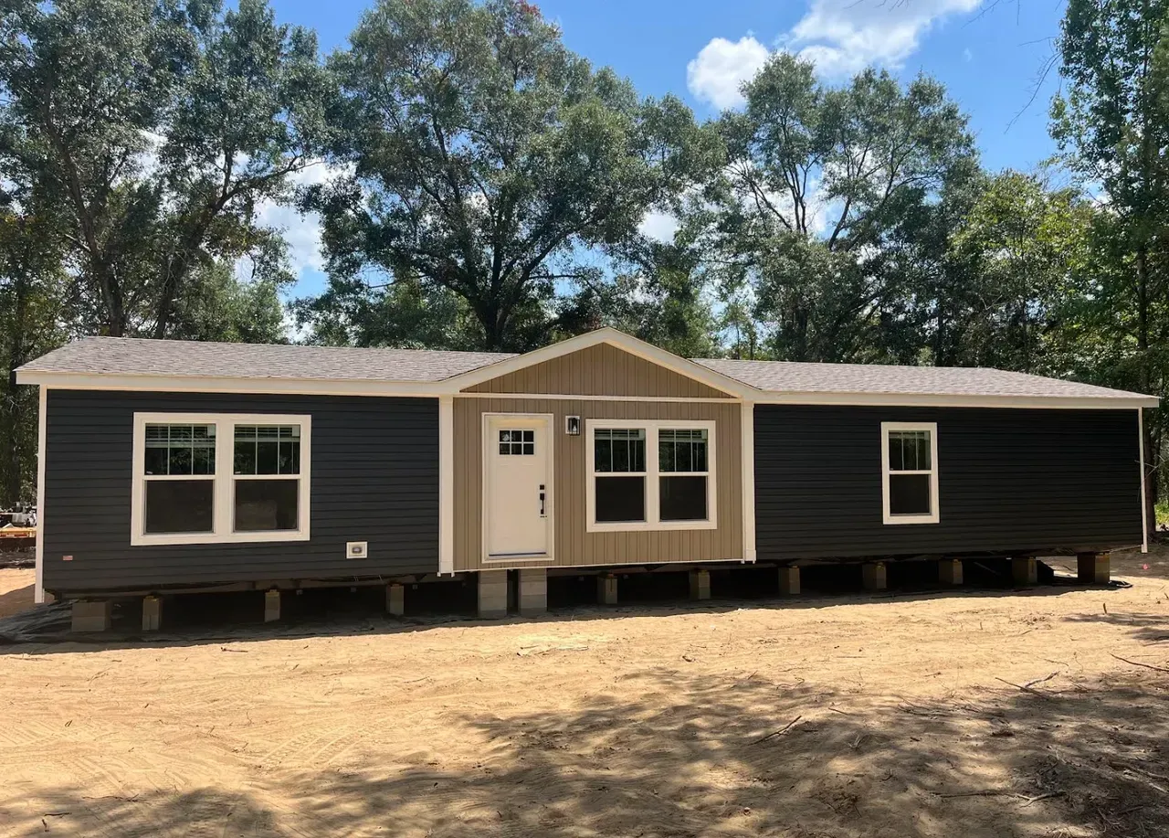 A single-story modular home with dark gray siding, a tan central entryway, and a shingled roof on a dirt lot.