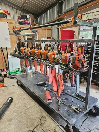 A cart holding numerous orange chainsaws with safety guards, in a cluttered workshop setting.