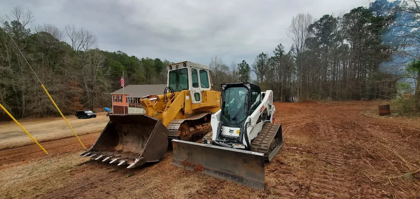 A bulldozer and skid steer on a construction site. Dirt ground and trees in the background under an overcast sky.