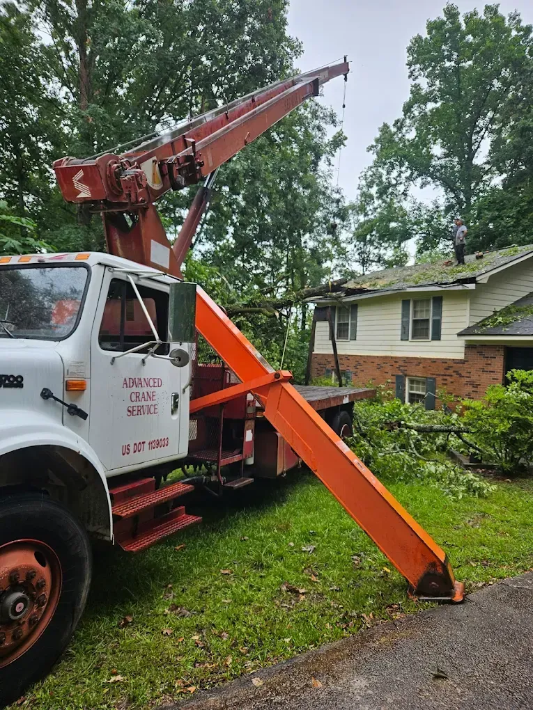 Truck with crane extended over house, likely for roof repair. Orange and white colors. Trees in background.