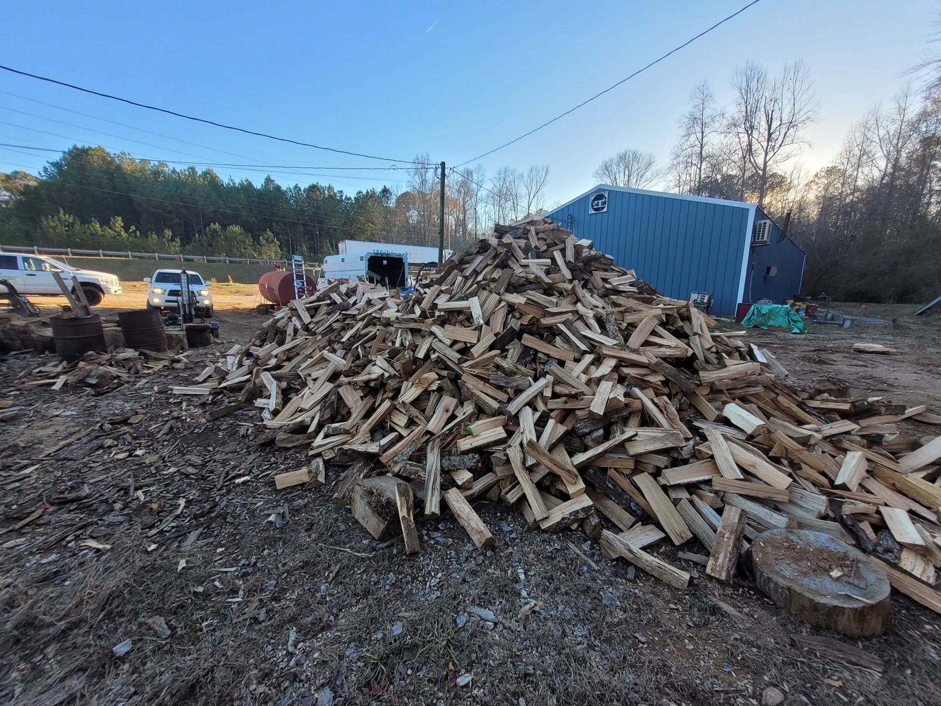 Pile of split firewood in a yard, with a blue building and a truck in the background. Sunny day.