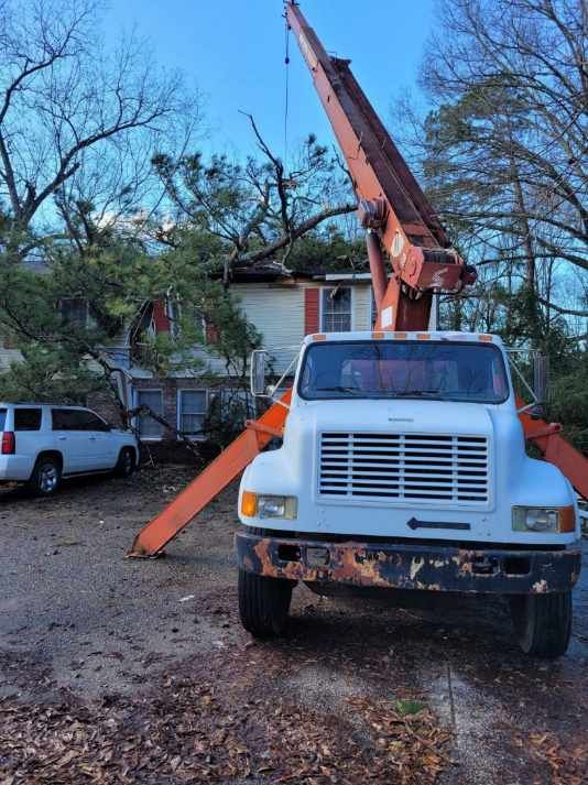 Crane truck removing a tree branch from a house. Orange boom extends upwards, white truck in front.