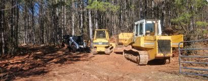 Three pieces of construction equipment on a dirt path, clearing land near a forest.