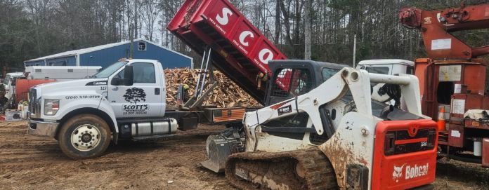 A white truck with a red dump bed being filled by a skid steer with firewood.