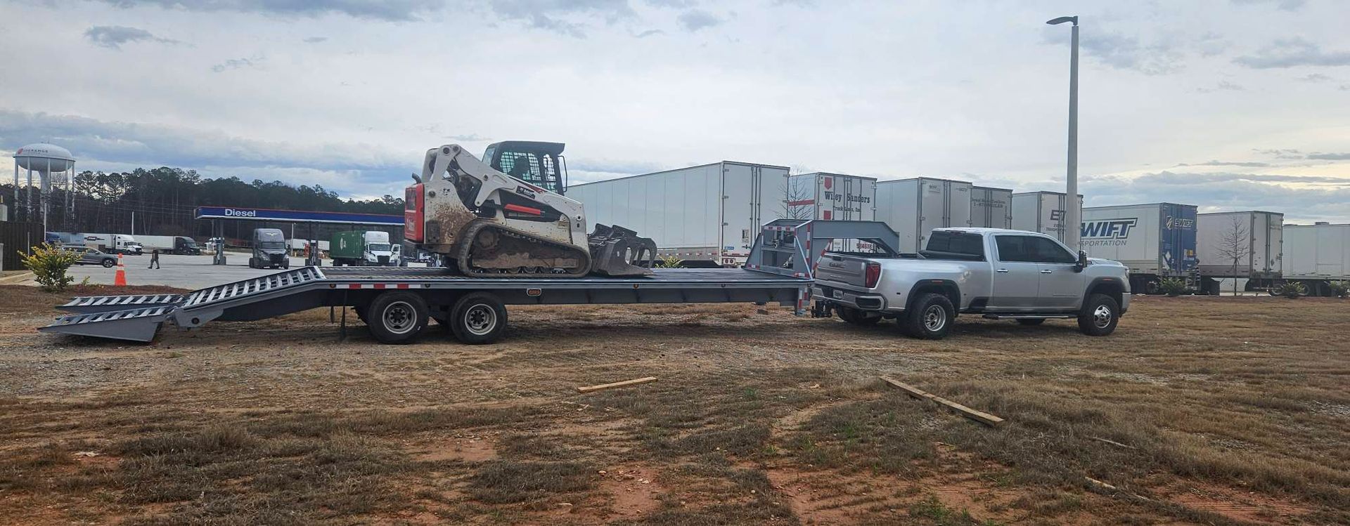 A silver pickup truck towing a flatbed trailer with a skid steer loader parked on it.