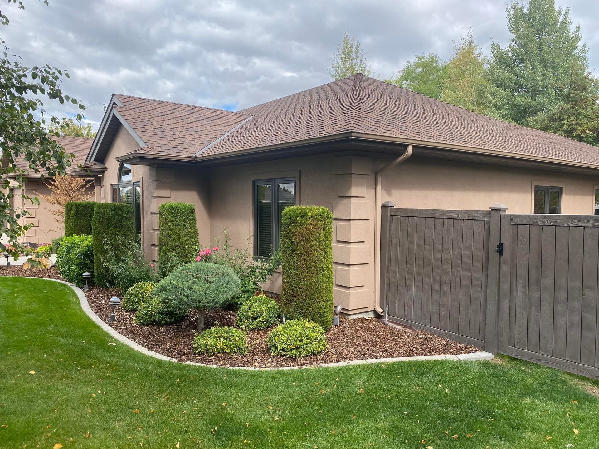 Beige house with a brown shingled roof, landscaping, and a wooden fence.