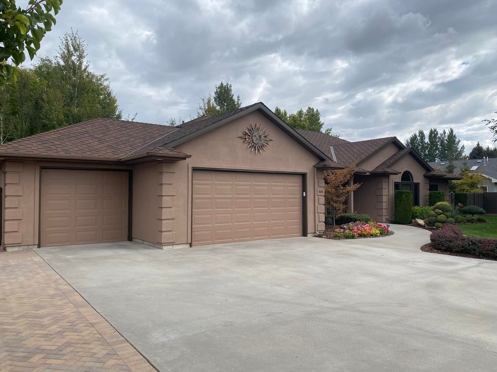 Tan stucco house with two-car garage, brown roof, and concrete driveway under cloudy sky.