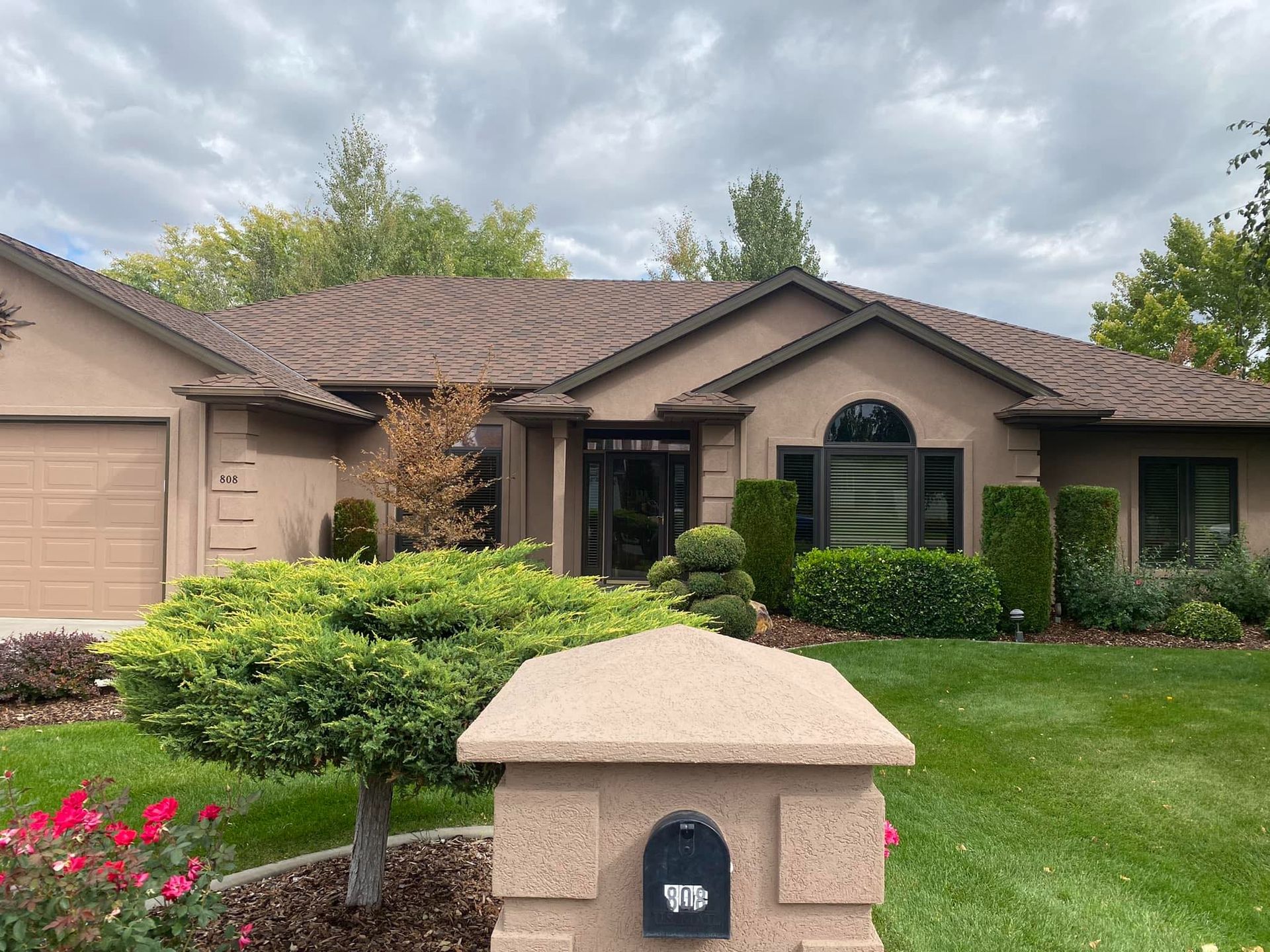 Tan stucco house with brown roof, green lawn, mailbox, and cloudy sky.