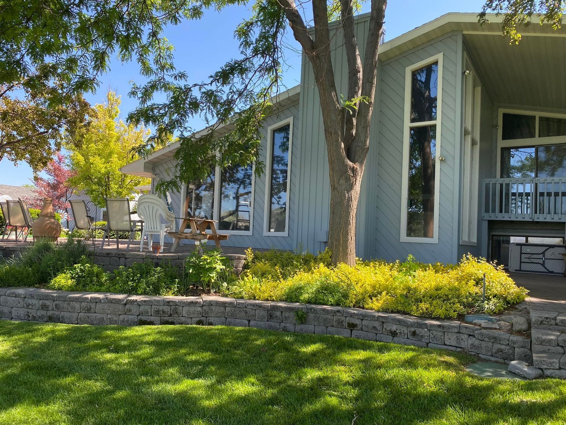 Blue house with tall windows, a retaining wall, lawn, and yellow plants. Trees with autumn colors visible.