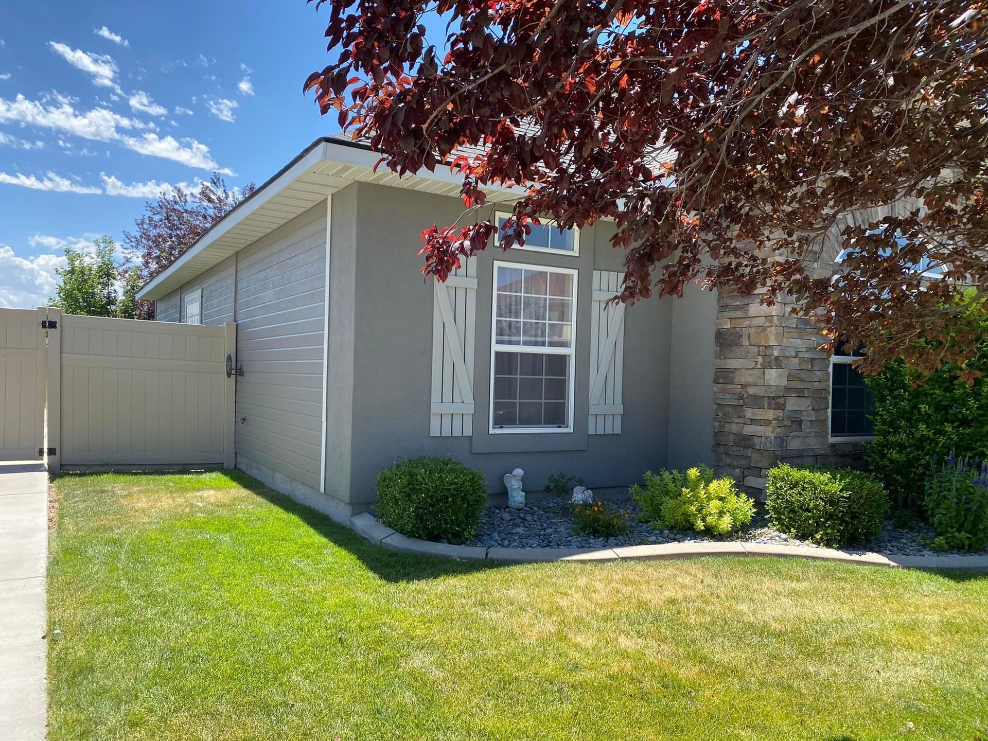 A house with light gray stucco and stone accents, shutters, and a beige fence, with lush green grass.