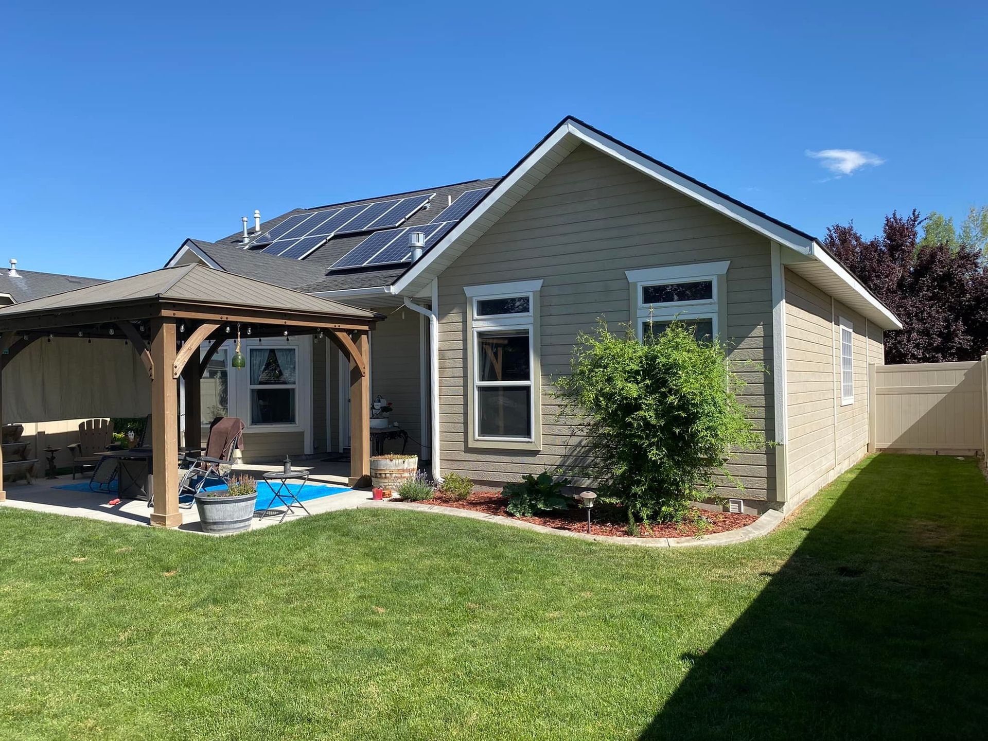 Backyard with a gazebo and house with solar panels on the roof. Green grass and blue sky.