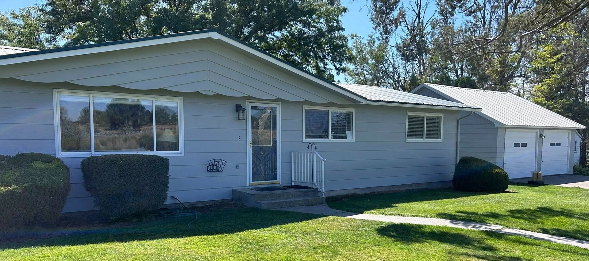 Gray house with a front porch, bushes, and a two-car garage under a sunny blue sky.