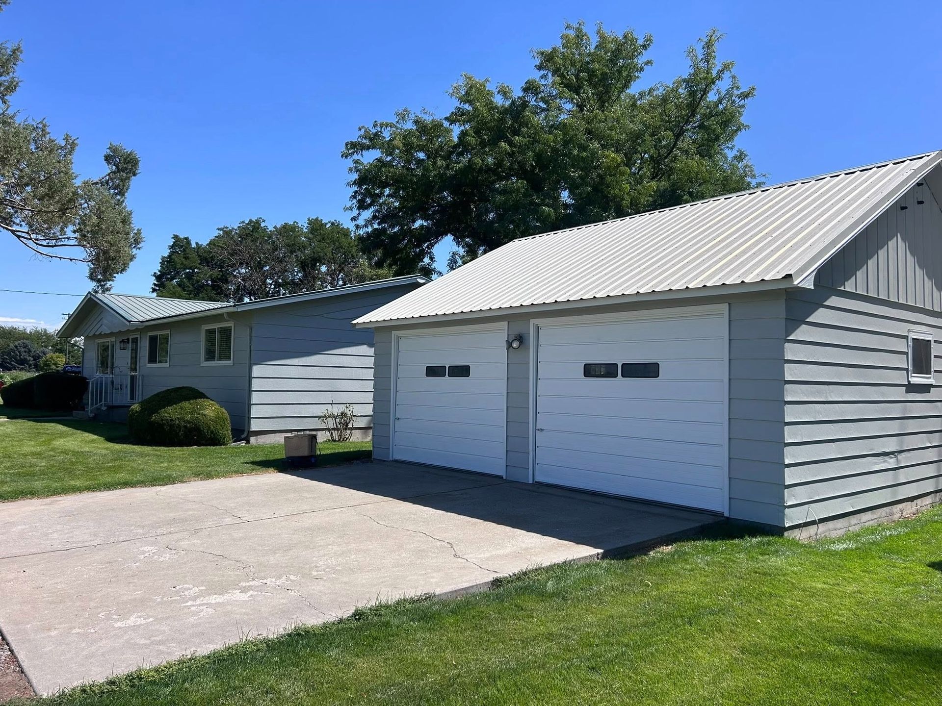 House with attached garage, both with light gray siding and metal roofs, on a sunny day. Concrete driveway and green lawn.
