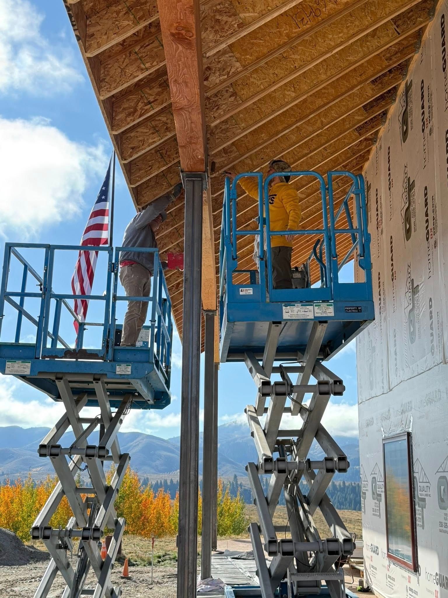 Two workers on scissor lifts install a building's roof under a blue sky, with an American flag.