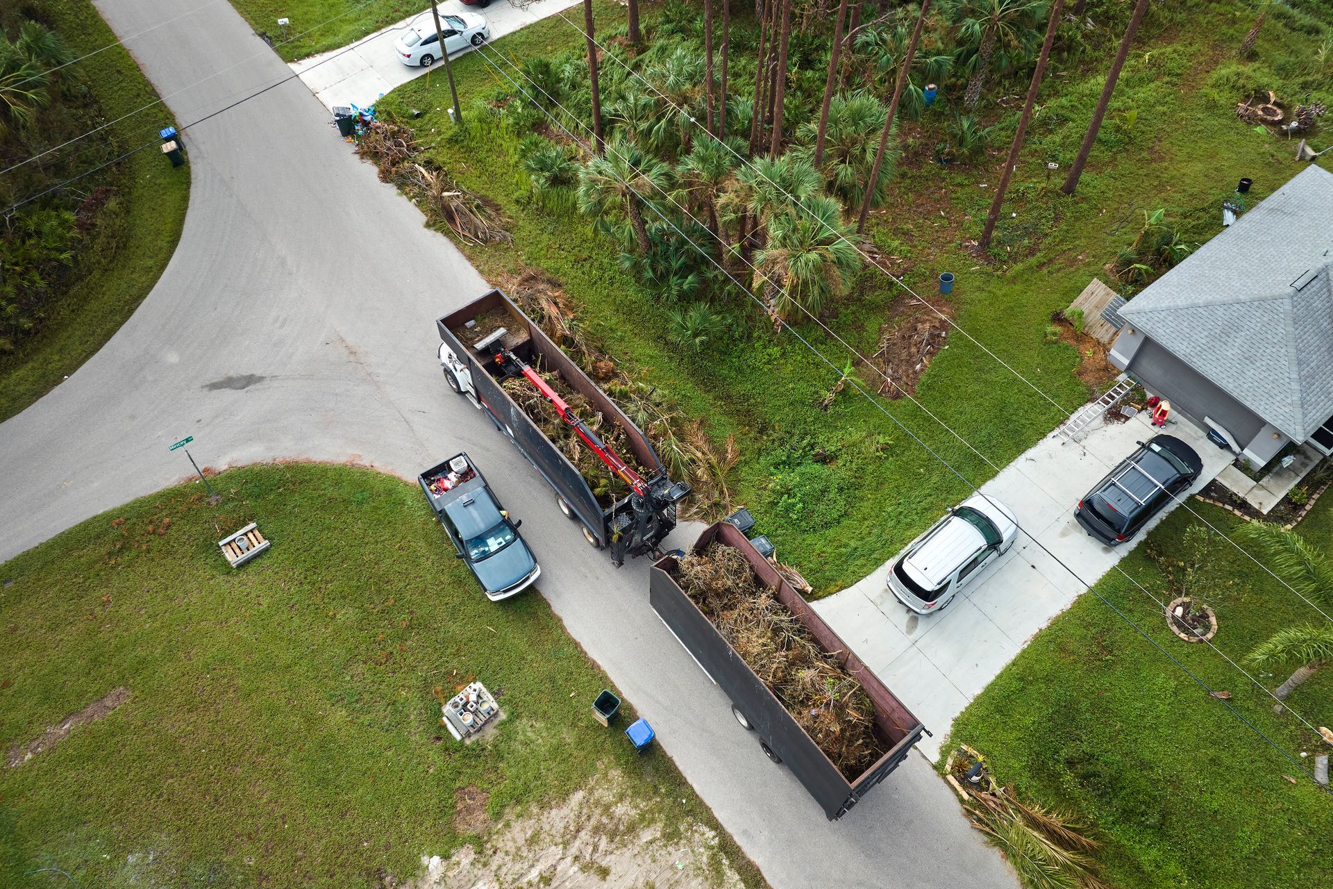 Aerial view of two large waste dumpsters parked along a suburban street with nearby houses and parked cars.