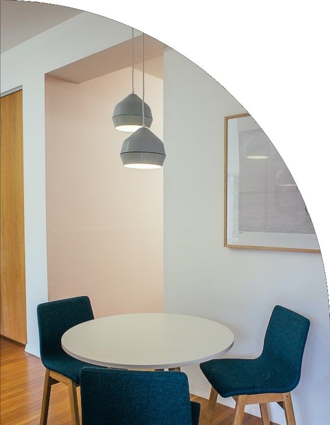 Round white table with three teal chairs under pendant lights in a dining area.