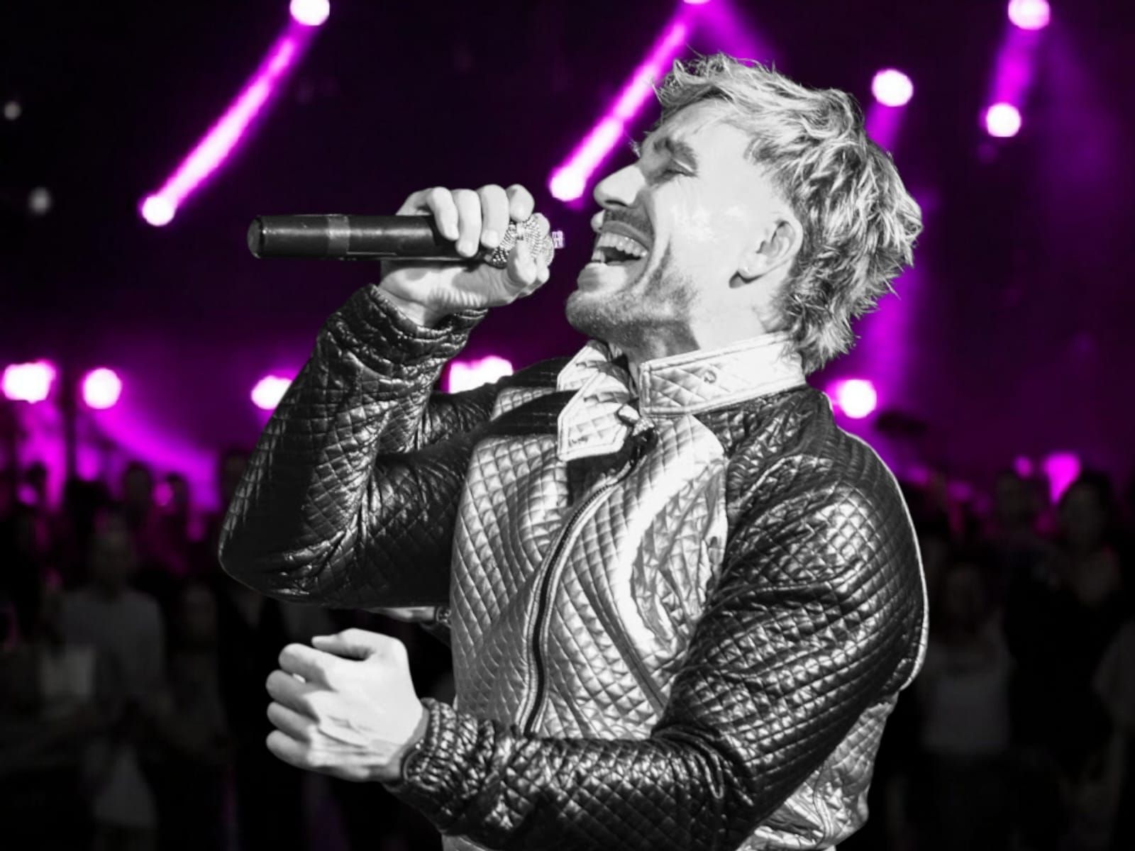 Cantante actuando en el escenario con un micrófono, con un traje blanco y negro contra luces de escenario moradas.
