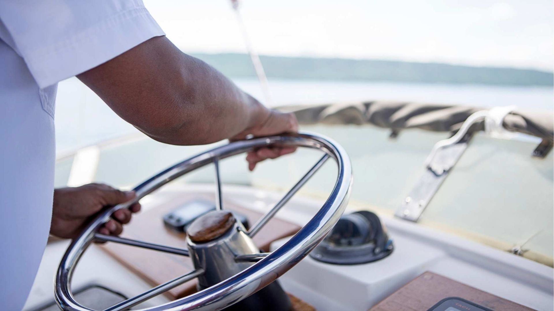 Person's hands on a boat's steering wheel, navigating a sunny body of water.