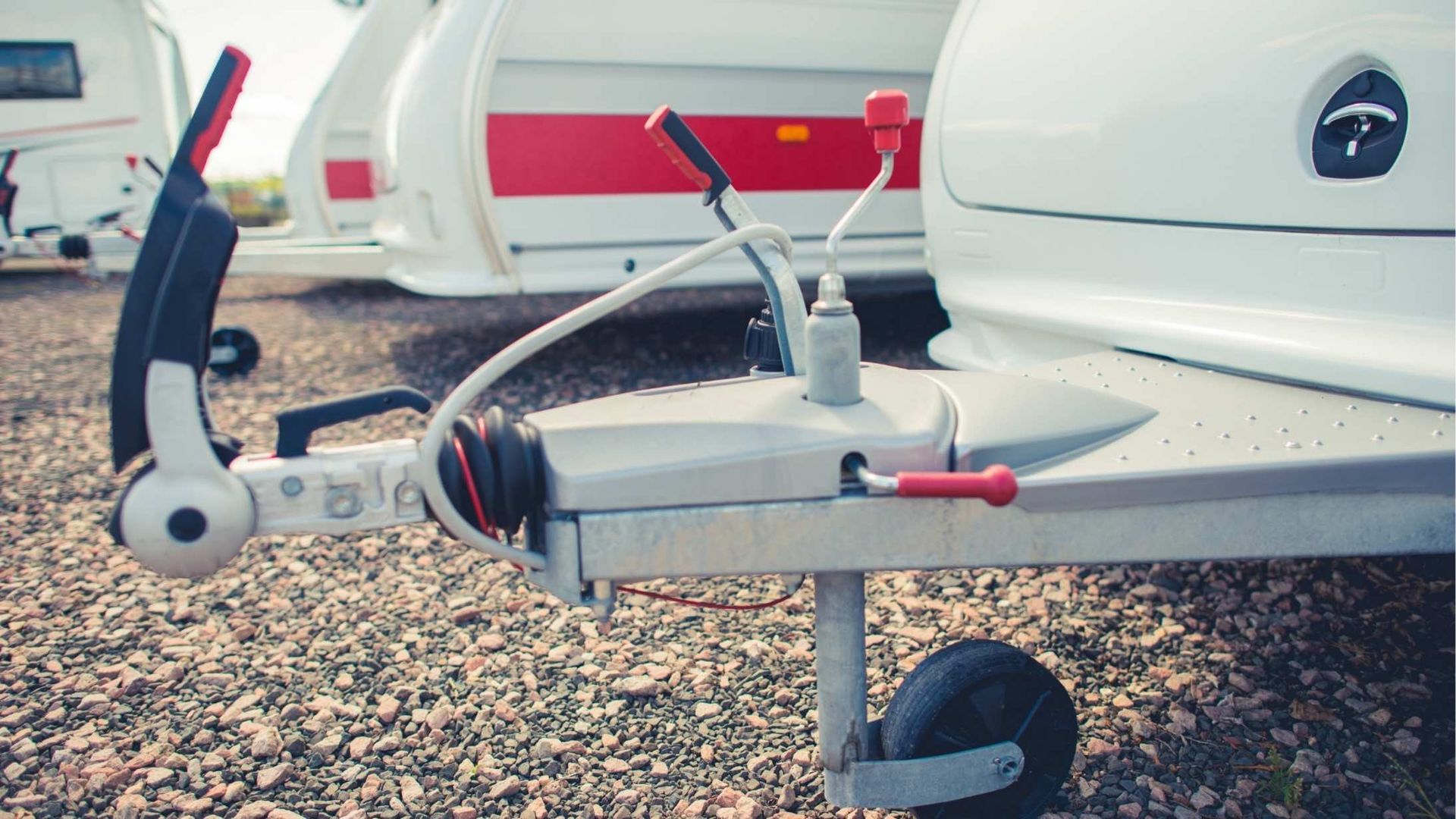 Close-up of a white trailer hitch, with levers and a stabilizing wheel, outdoors on gravel.