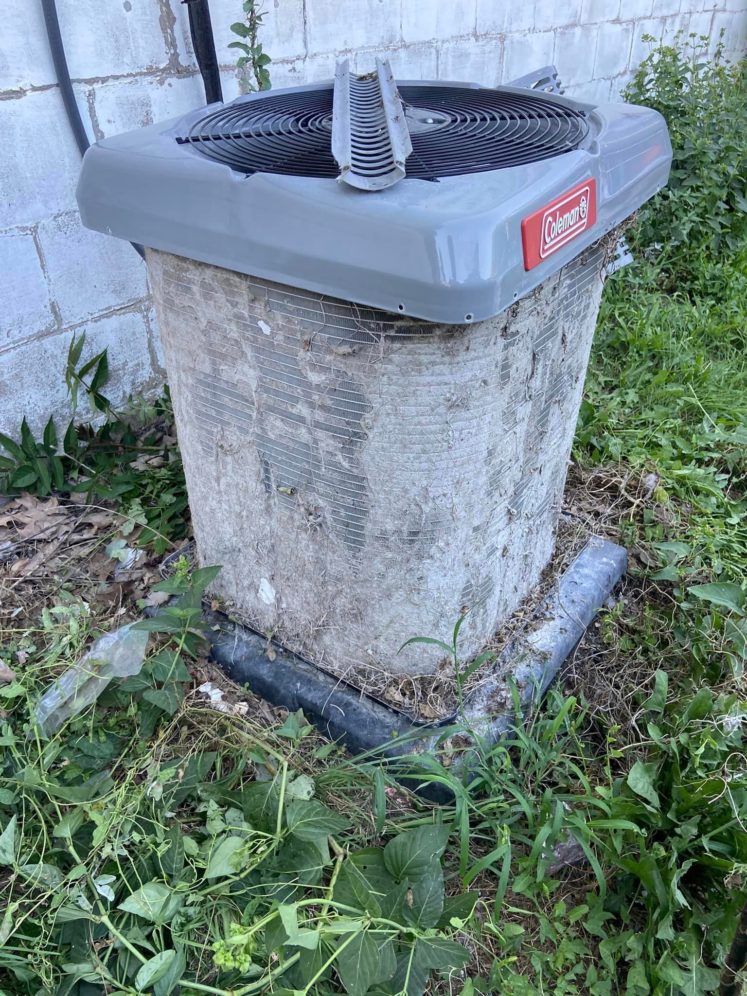 A dirty air conditioner is sitting in the grass next to a brick wall.