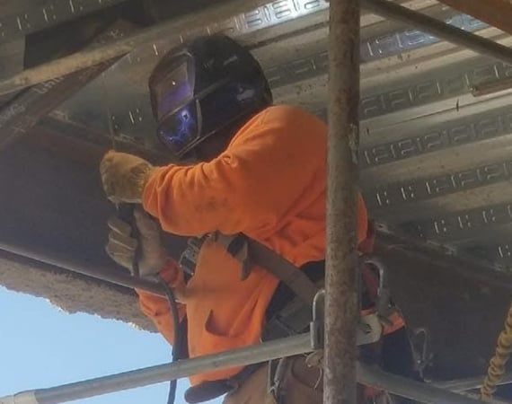 A man wearing a welding mask is standing on a scaffolding