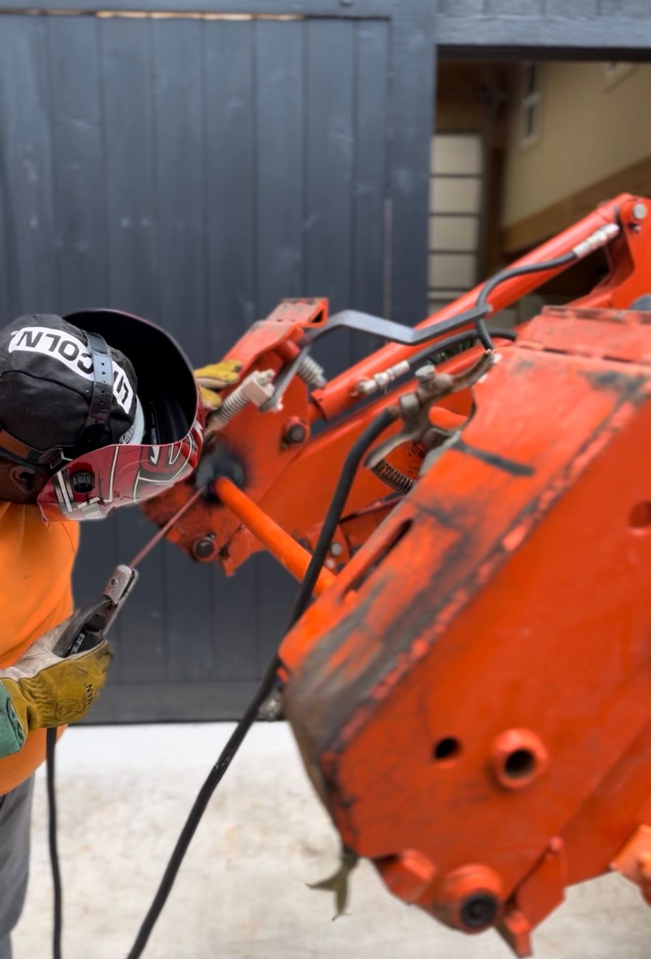 A man wearing a welding helmet is working on an orange machine.