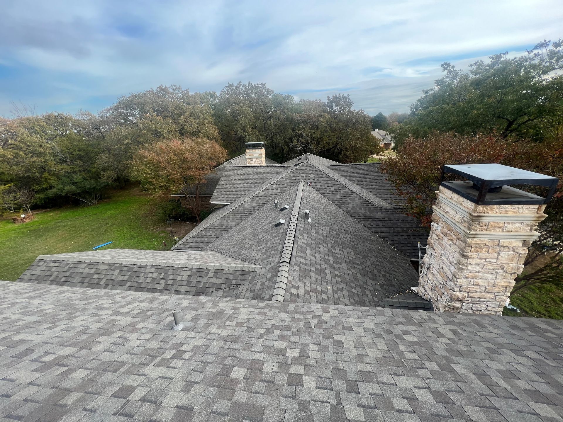 View of a shingled roof with chimneys and trees against a cloudy sky.