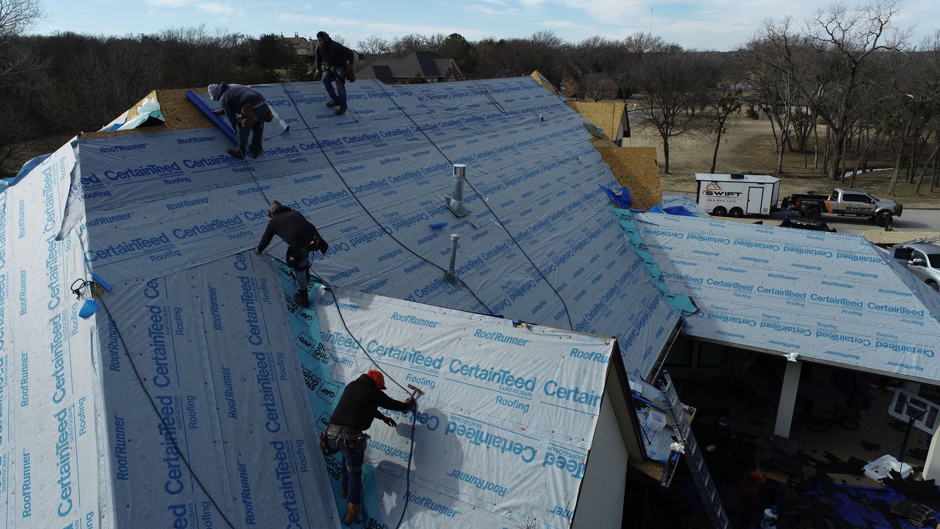 Roofers installing blue underlayment on a multi-gabled roof, sunny day.