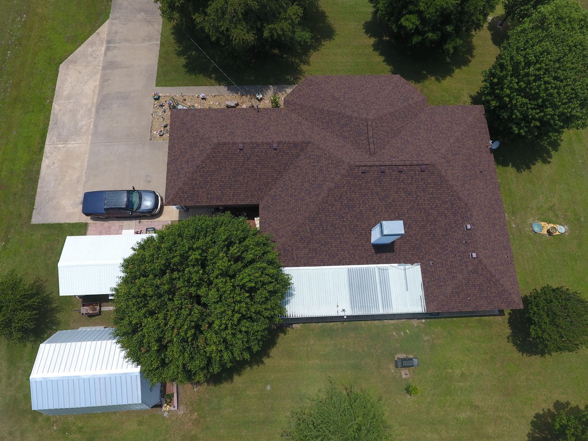 An aerial view of a house with a dark brown shingled roof, an attached white carport, a driveway, and surrounding trees.