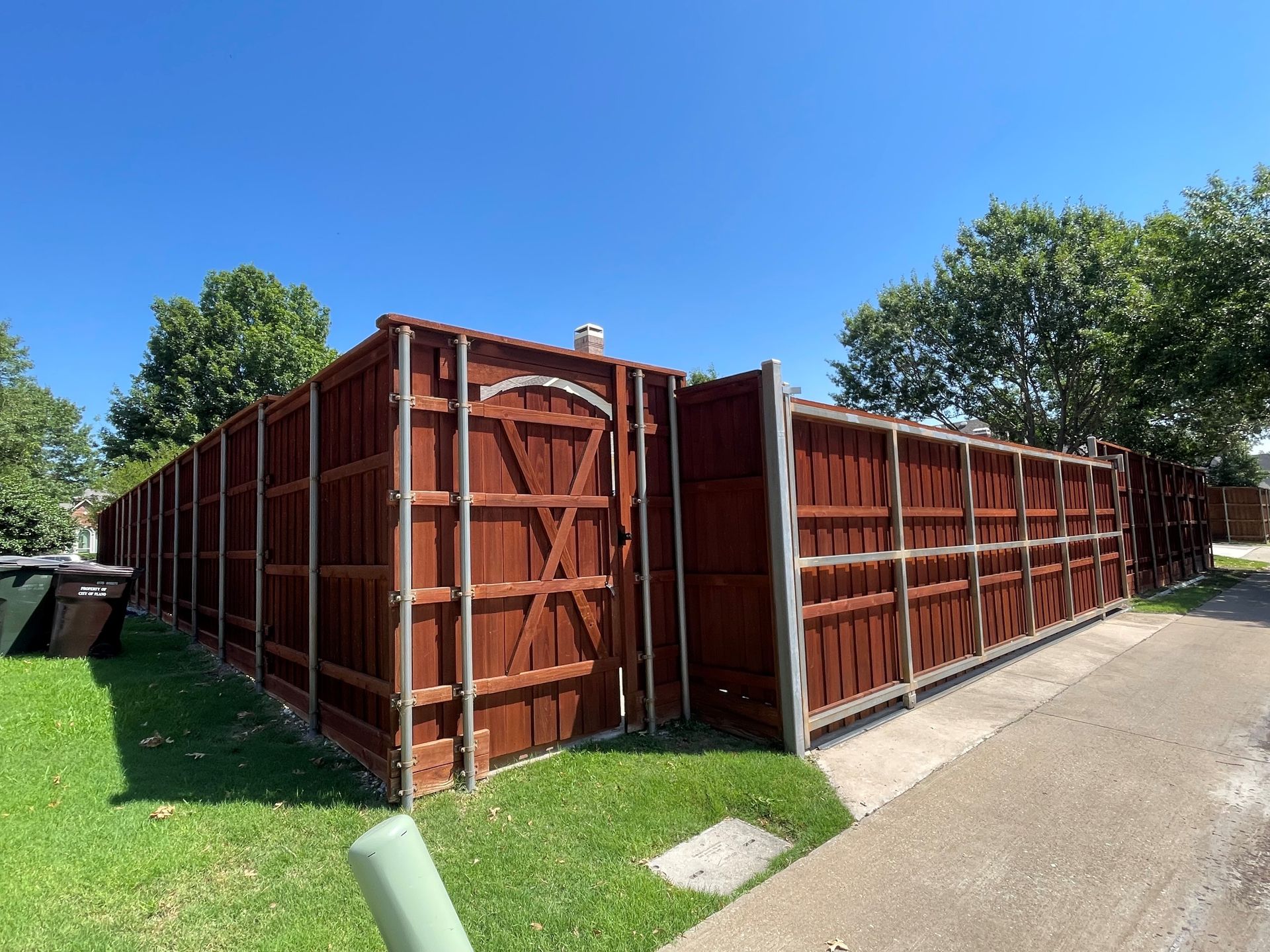 A tall, stained brown wooden privacy fence with a gated entrance sits on a grassy lawn next to a concrete sidewalk.