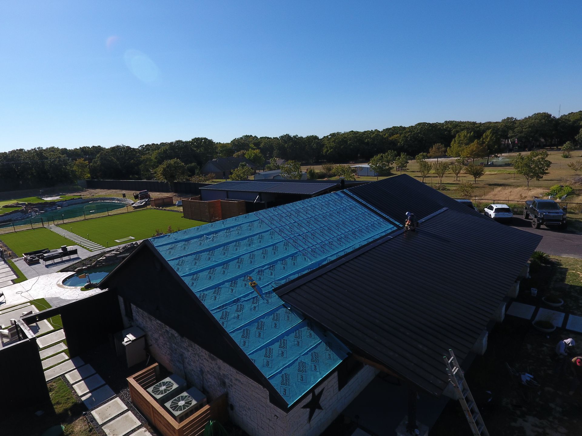 An aerial view of a house roof partially covered with blue underlayment and partially finished with black metal roofing.