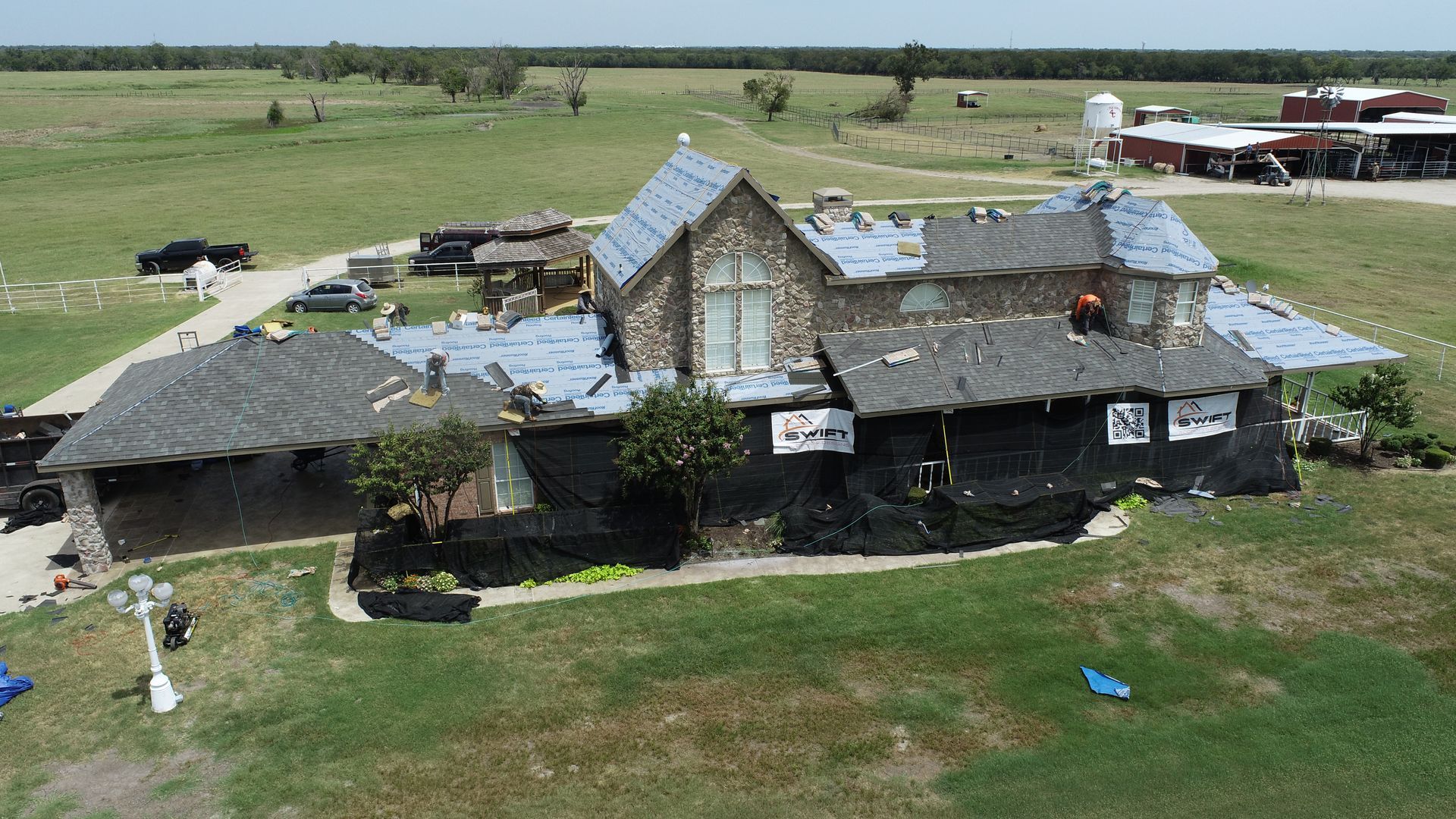 Drone view of a stone house with roof repairs, surrounded by green fields and a barn in the distance.