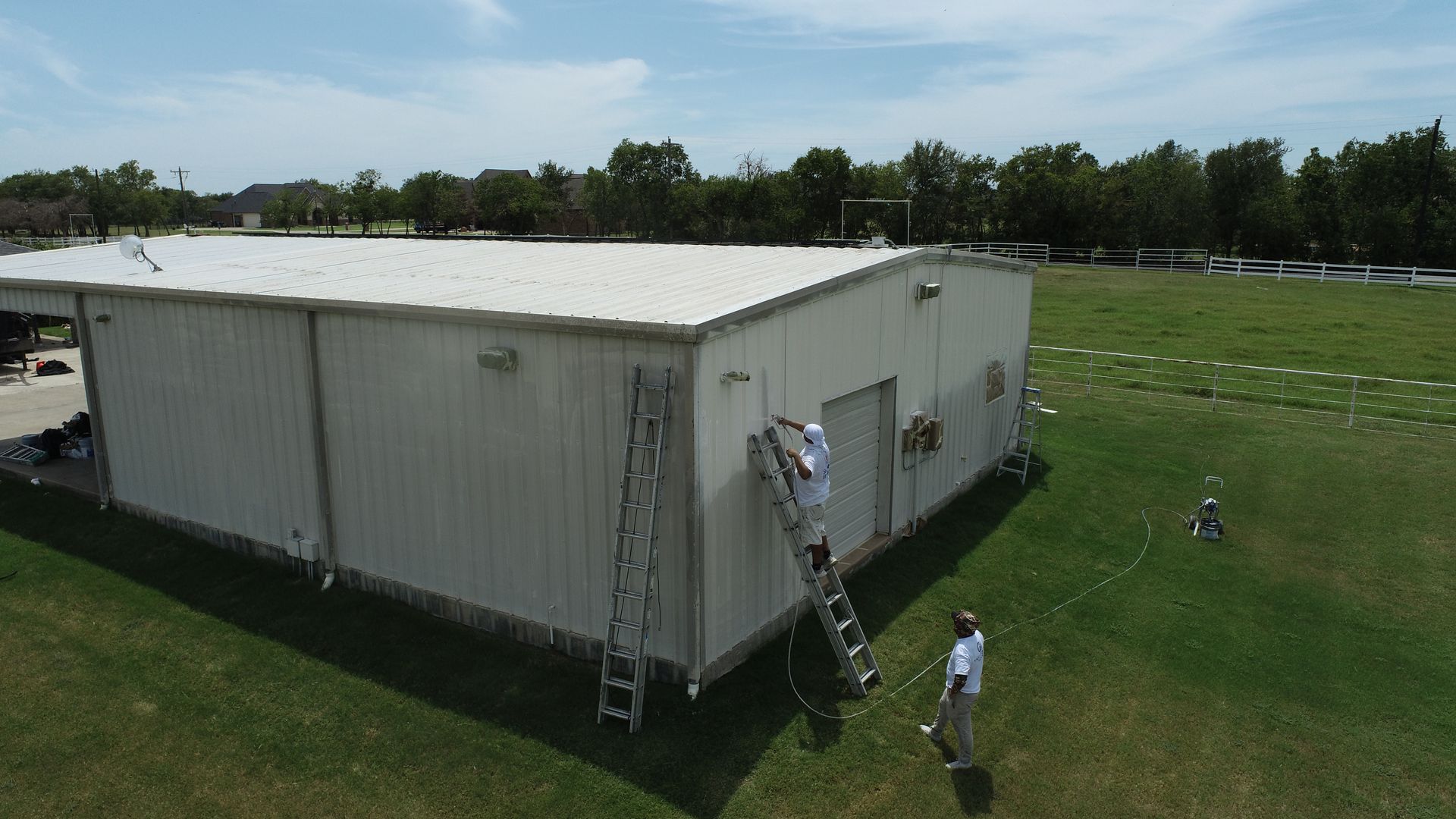 Two people painting a white metal building on a grassy field. One is on a ladder.