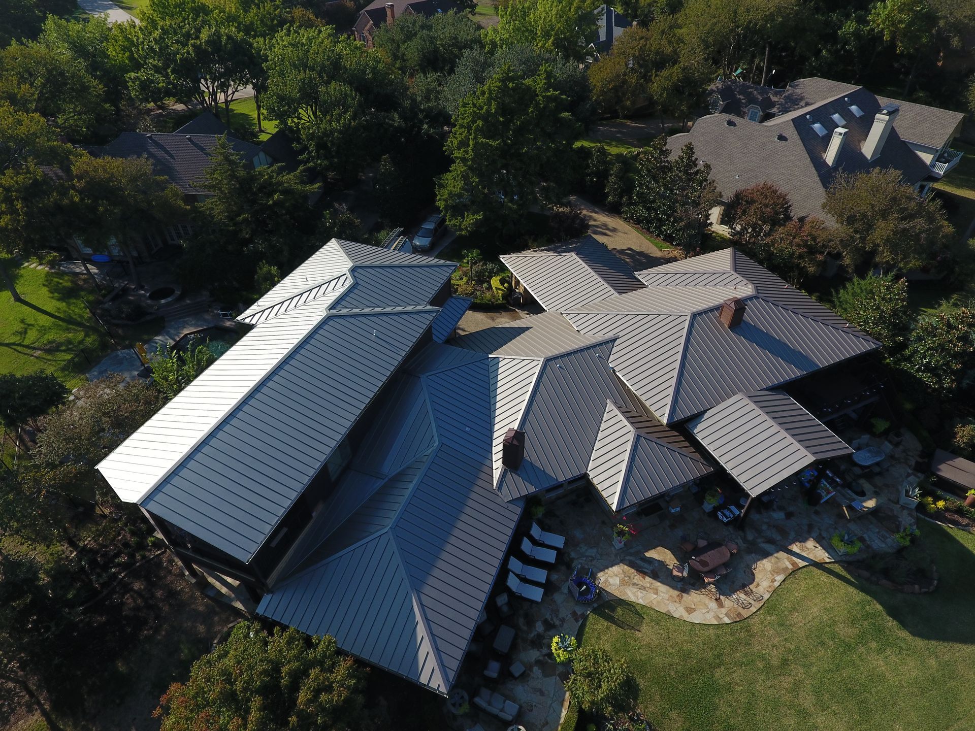 Aerial view of a large house with a complex, gray metal roof, surrounded by trees and a green lawn.
