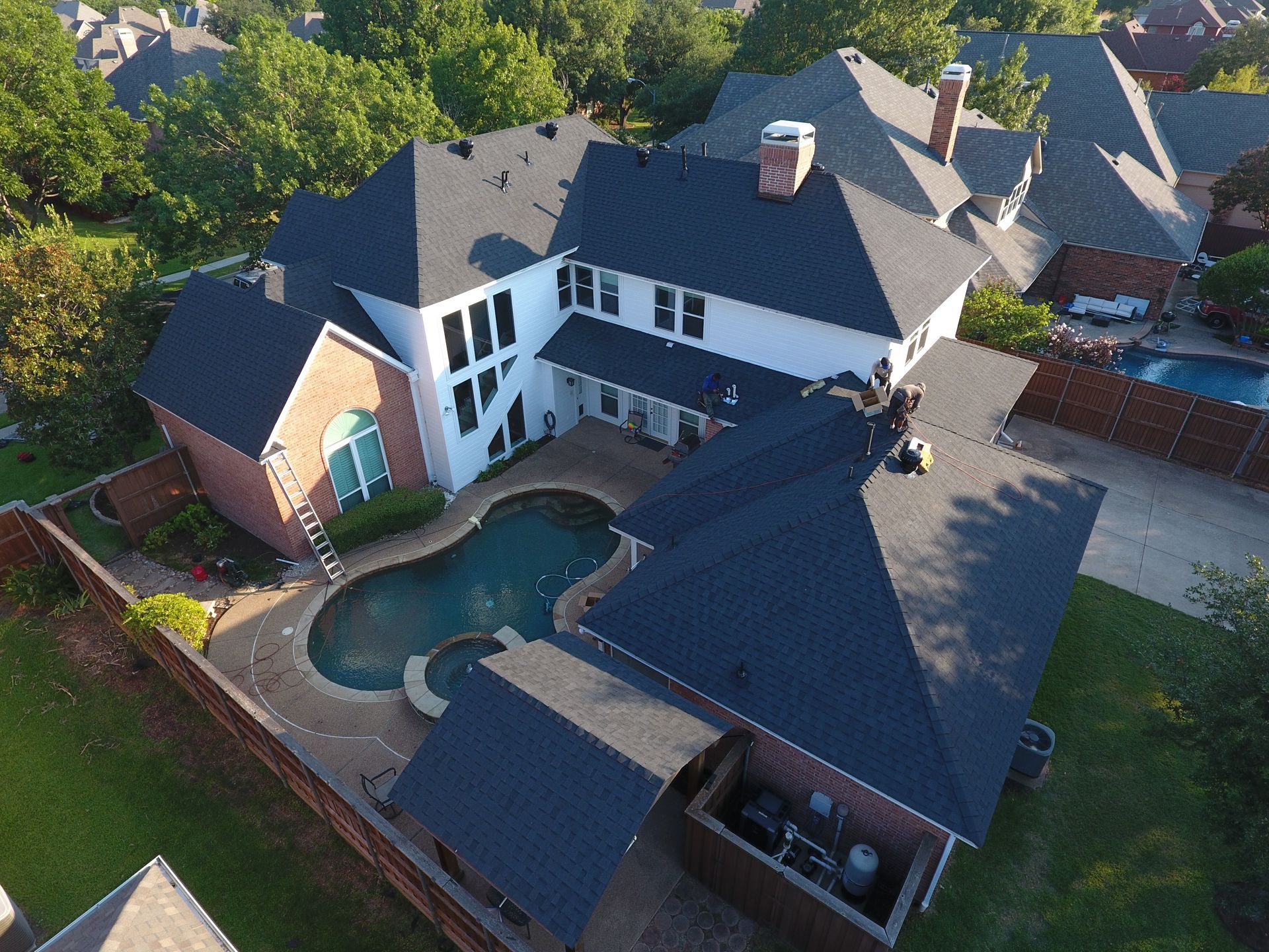 Aerial view of a large house with a pool and dark roof surrounded by trees.