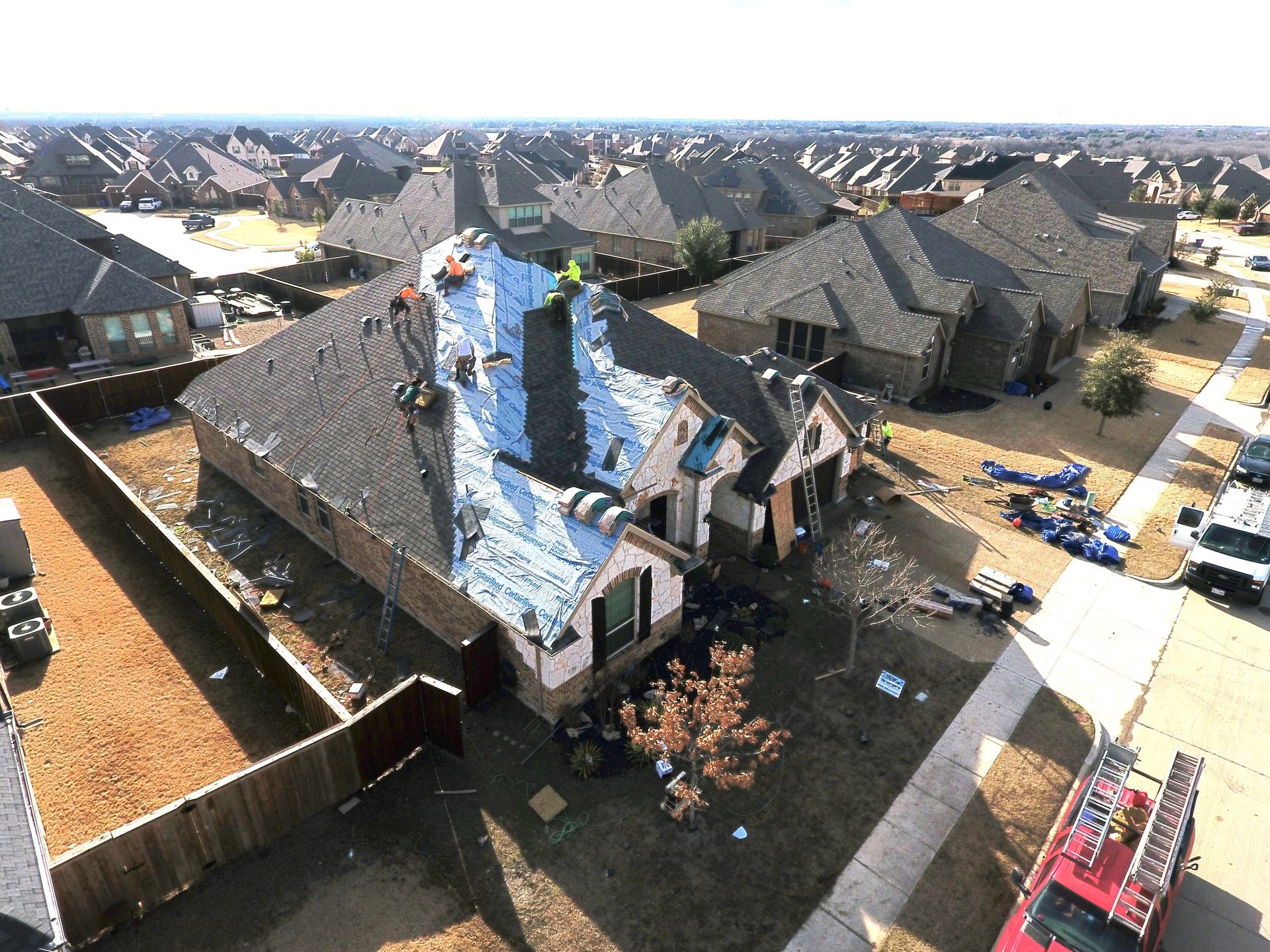 Aerial view of a house being re-roofed with workers on top. Other homes surround it in a suburban neighborhood.