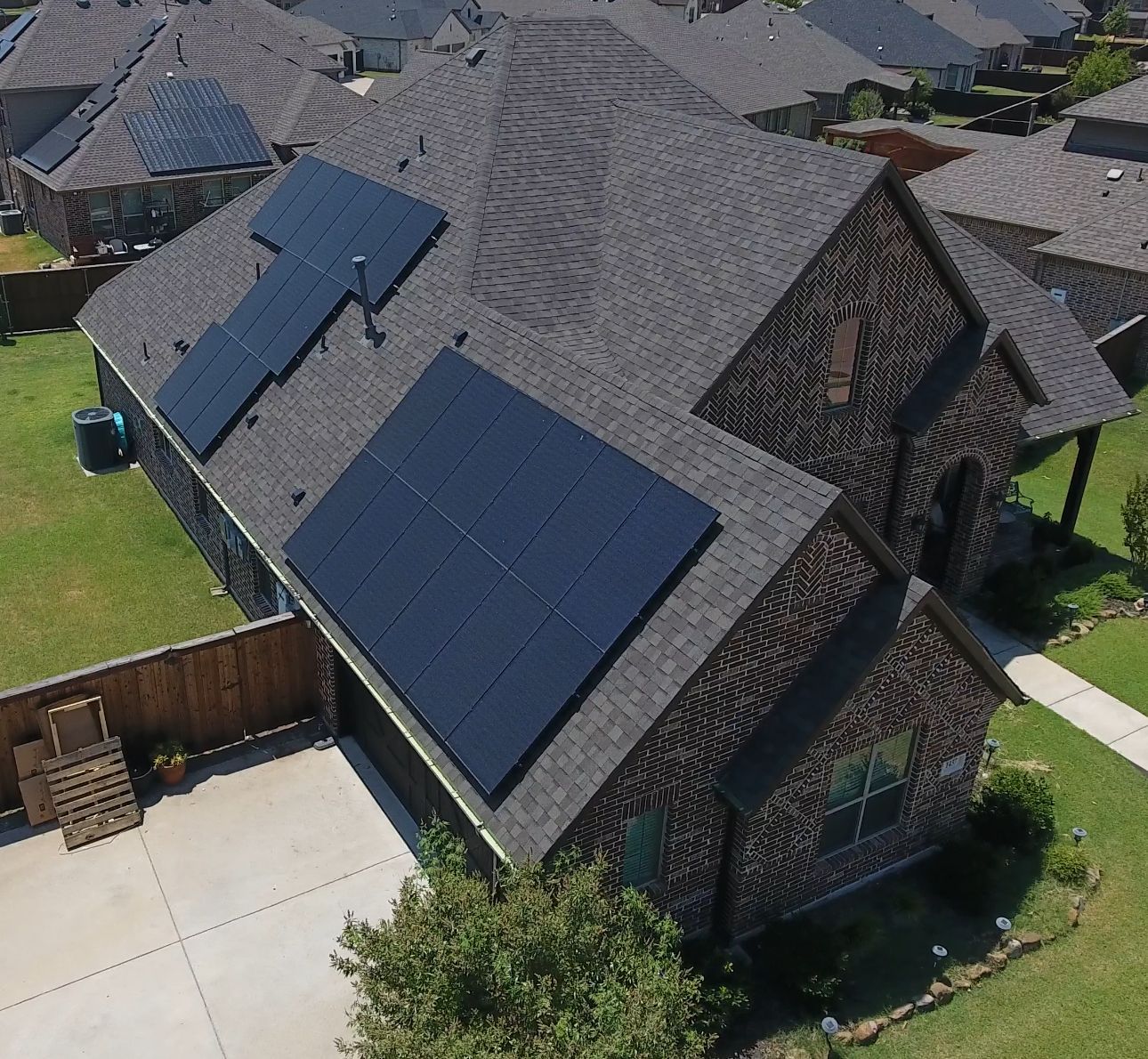 Solar panels on a dark-shingled roof of a brick house in a suburban neighborhood.
