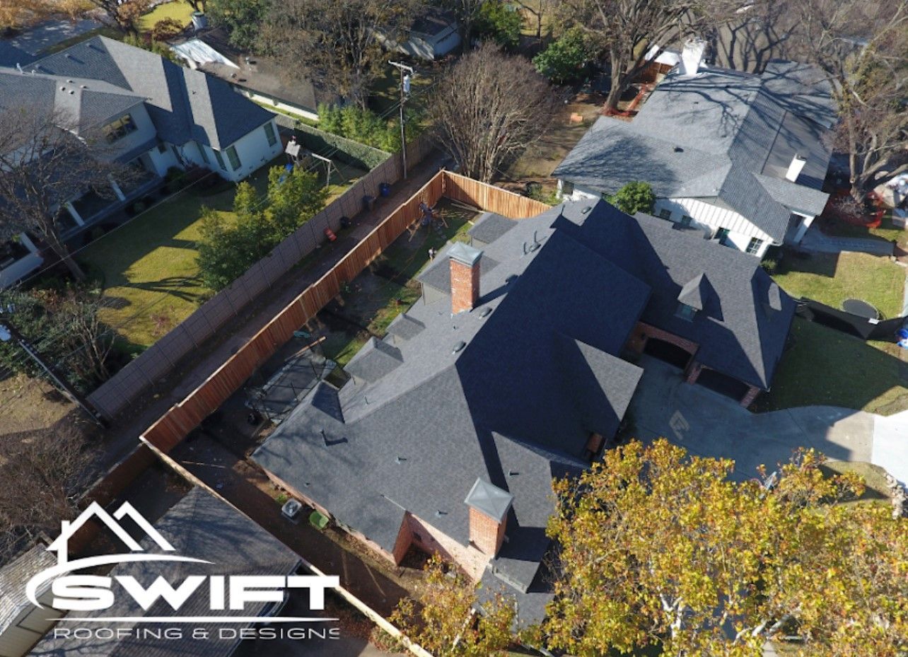 Aerial view of a dark gray shingled roof on a house, with a chimney, surrounded by green grass, fence, and other houses.