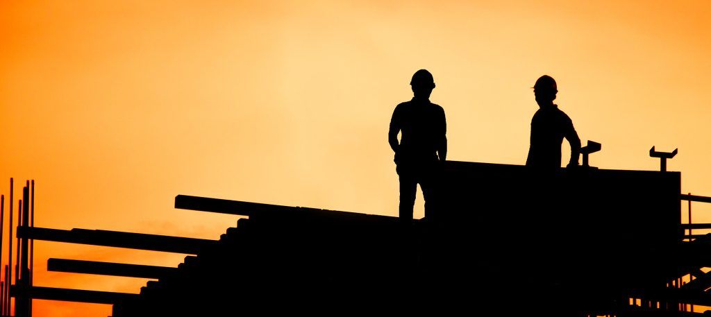Two construction workers are standing on top of a building at sunset.