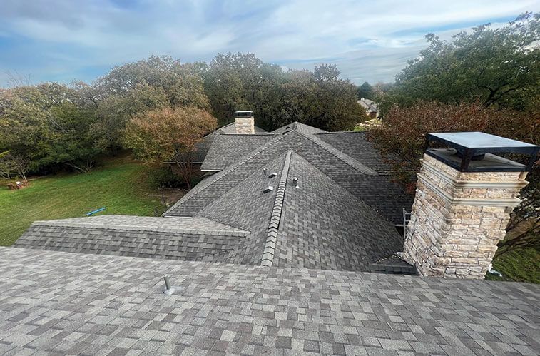 Gray asphalt shingle roof on a house, stone chimney, surrounded by trees and lawn.