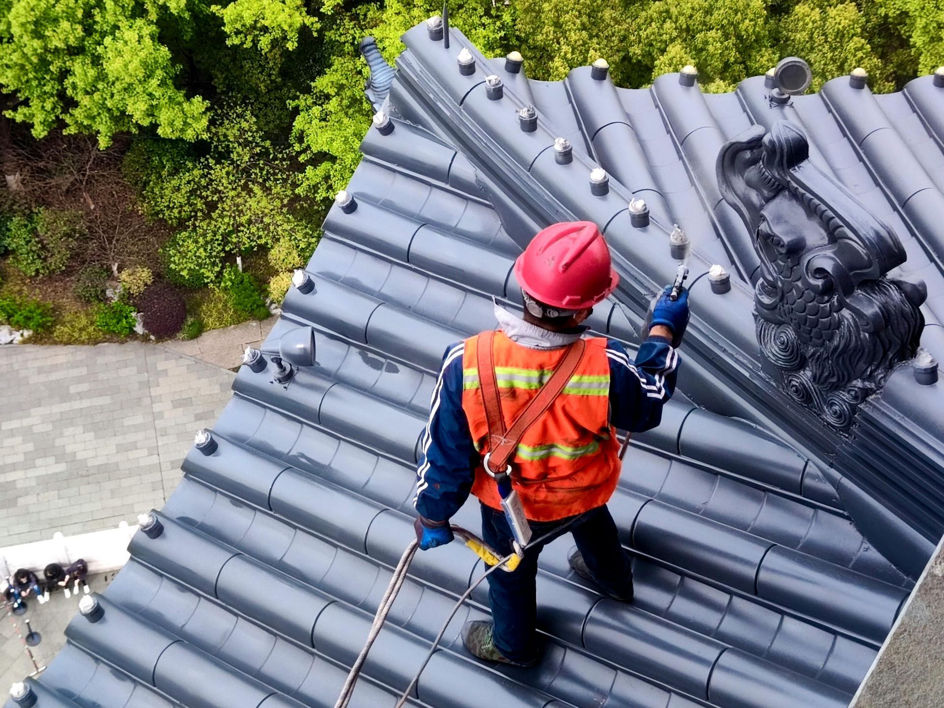 Roofer in safety gear on a dark tiled roof with a dragon sculpture, green trees in the background.