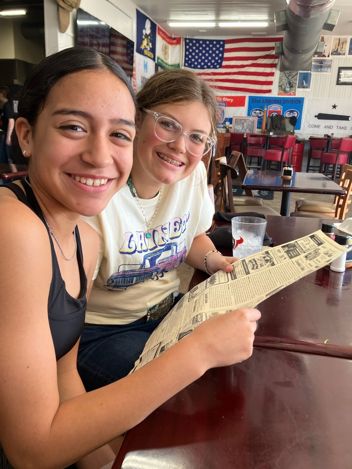 Two smiling people seated at a table, looking at a menu in a diner; American flag in the background.
