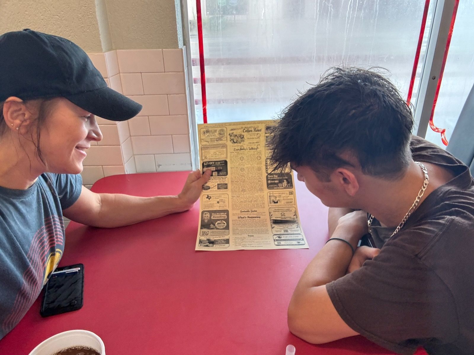 Two people sit at a red table, looking at a menu. One wears a black cap and smiles. The other has short dark hair.