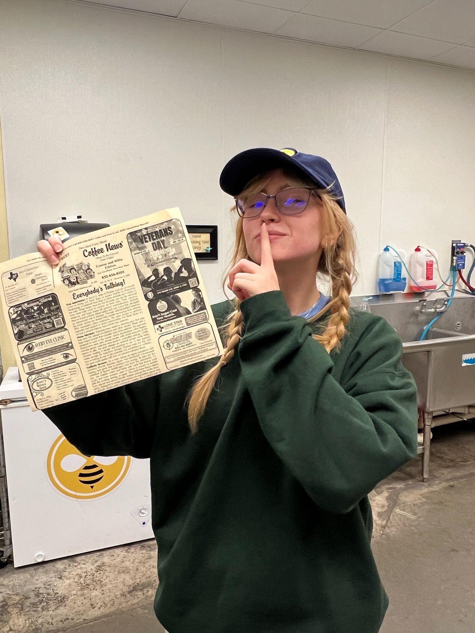 Person in a green sweatshirt holding old paper, finger to lips, wearing a hat in a kitchen area.