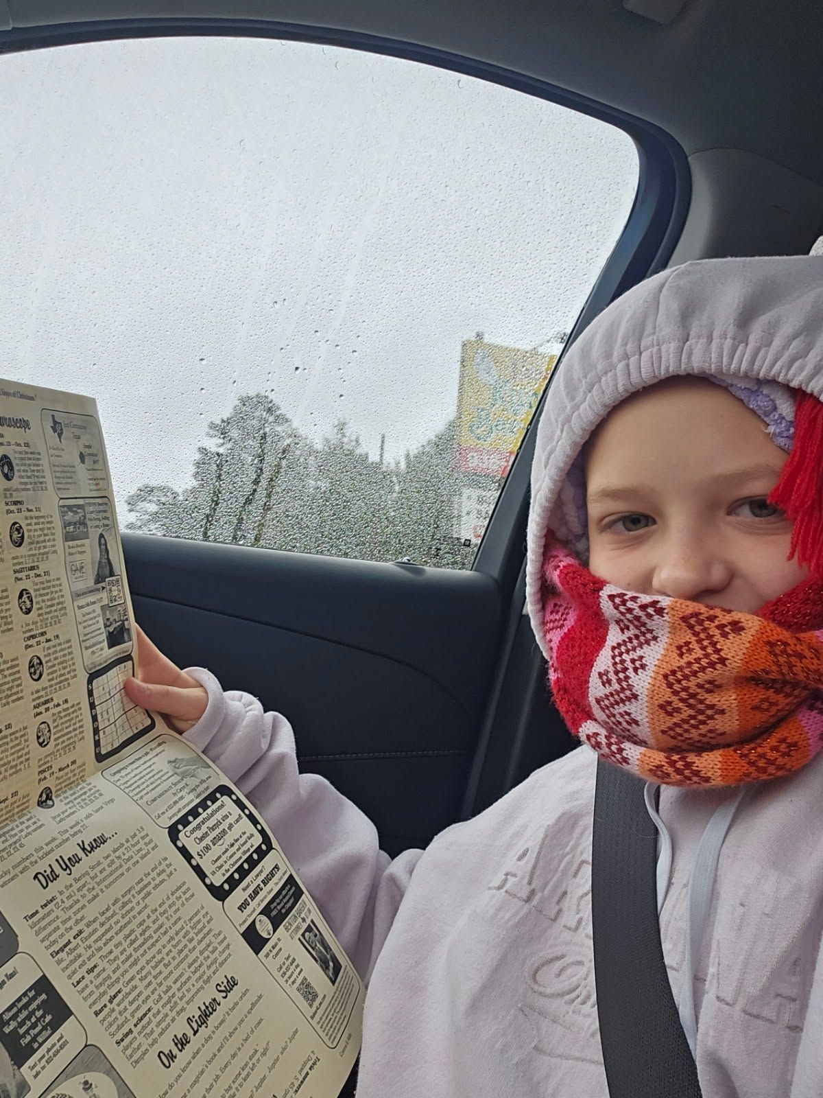 Girl in a car, reading a newspaper with raindrops on the window. She is wearing a hooded jacket and scarf.