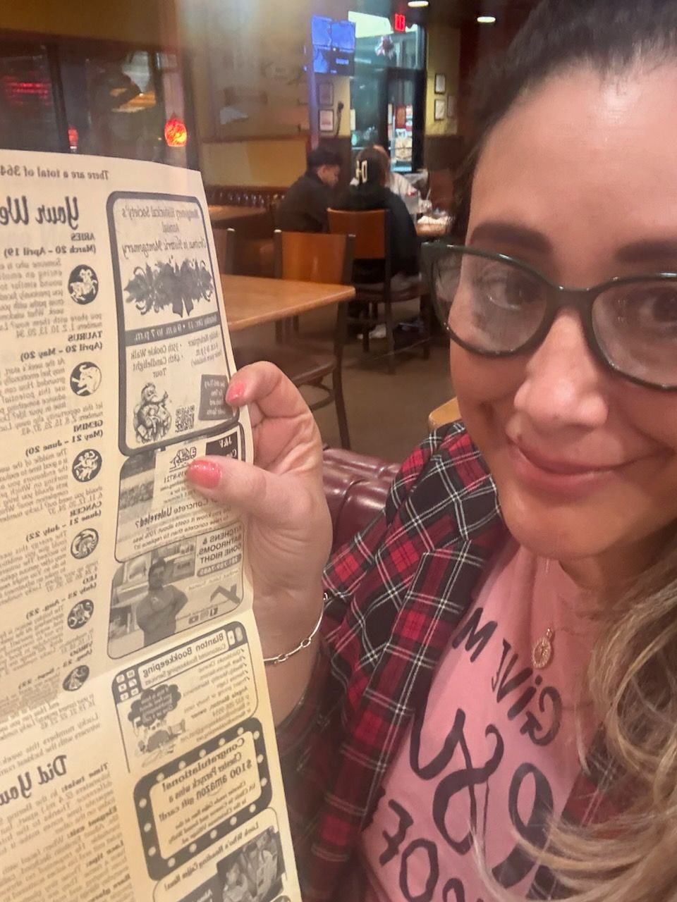 Woman in glasses holding a newspaper in a restaurant, smiling. Red and black plaid shirt, pink shirt.
