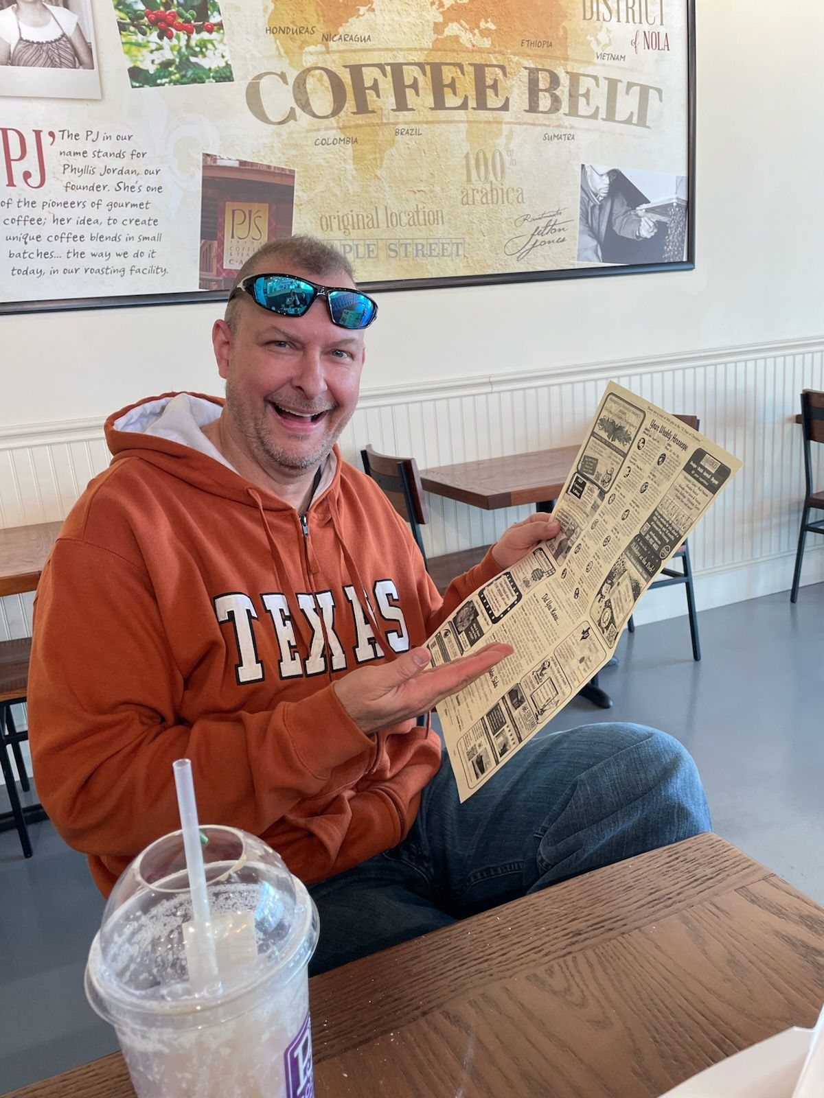 Man in orange Texas hoodie smiles, holding a newspaper, seated in a coffee shop.