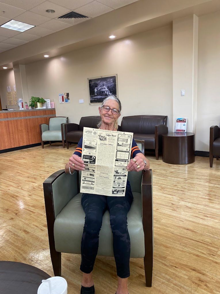 Woman in chair holding newspaper; sitting in a waiting room with tan walls and hardwood floors.
