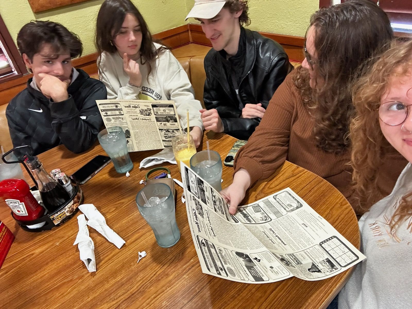 Five people at a restaurant table look at menus. Drinks and condiments are on the wooden table.